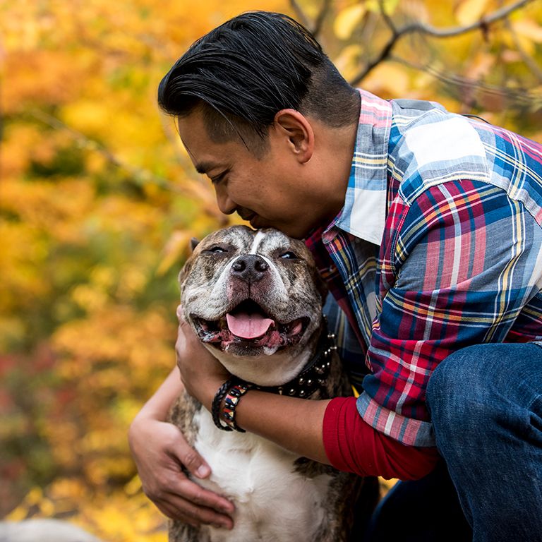 A man hugging a dog in front of trees with autumn colors